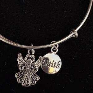 Puede incluir: Pulsera de brazalete plateada con dos colgantes. Uno es un &aacute;ngel detallado, el otro un colgante redondo con la palabra "Faith". La pulsera se muestra sobre un fondo negro.