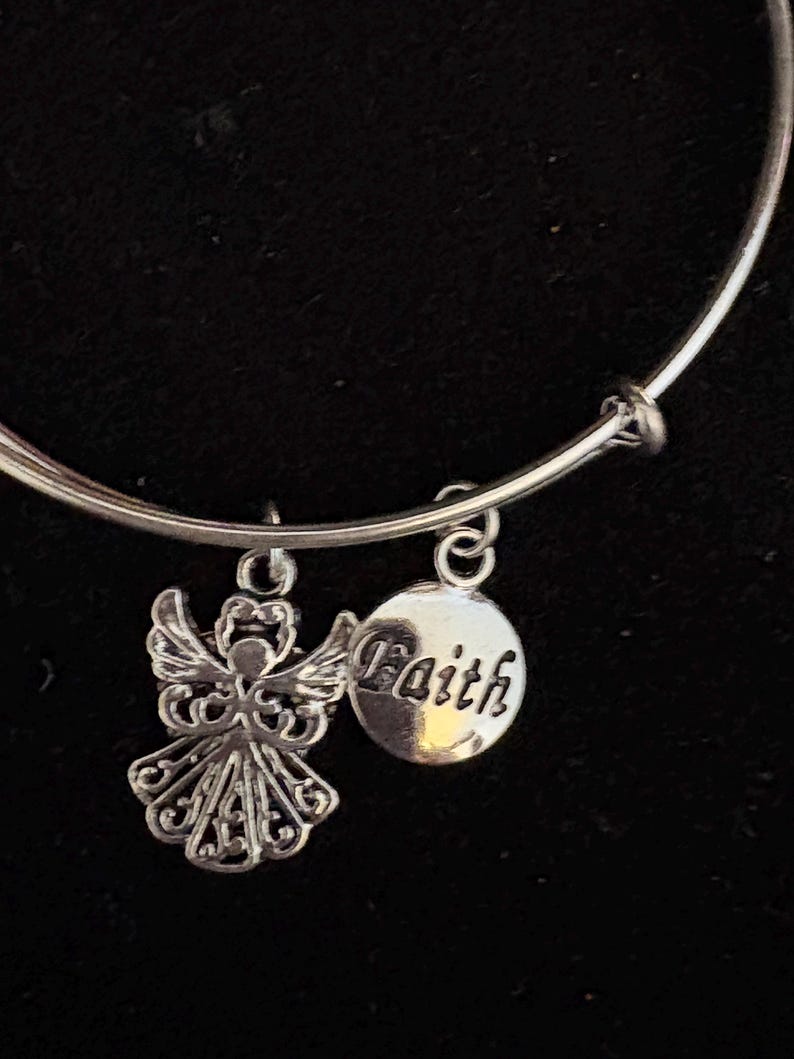 Puede incluir: Pulsera de brazalete plateada con dos colgantes. Uno es un &aacute;ngel detallado, el otro un colgante redondo con la palabra "Faith". La pulsera se muestra sobre un fondo negro.