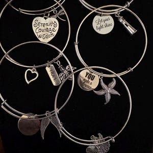 Puede incluir: Pulseras de dijes plateadas con varios colgantes. Los dijes incluyen un coraz&oacute;n, una lib&eacute;lula, alas, una estrella de mar y un faro. Algunos dijes tienen palabras inspiradoras como "Serenity, Courage, Wisdom" y "You make a difference".