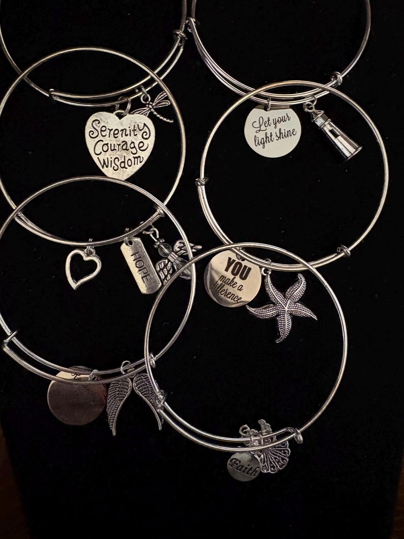Puede incluir: Pulseras de dijes plateadas con varios colgantes. Los dijes incluyen un coraz&oacute;n, una lib&eacute;lula, alas, una estrella de mar y un faro. Algunos dijes tienen palabras inspiradoras como "Serenity, Courage, Wisdom" y "You make a difference".