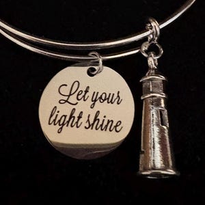 Puede incluir: Pulsera de brazalete plateada con un dije redondo y un dije de faro. El dije redondo tiene el texto "Let your light shine". El dije de faro tiene ventanas y un techo detallados.