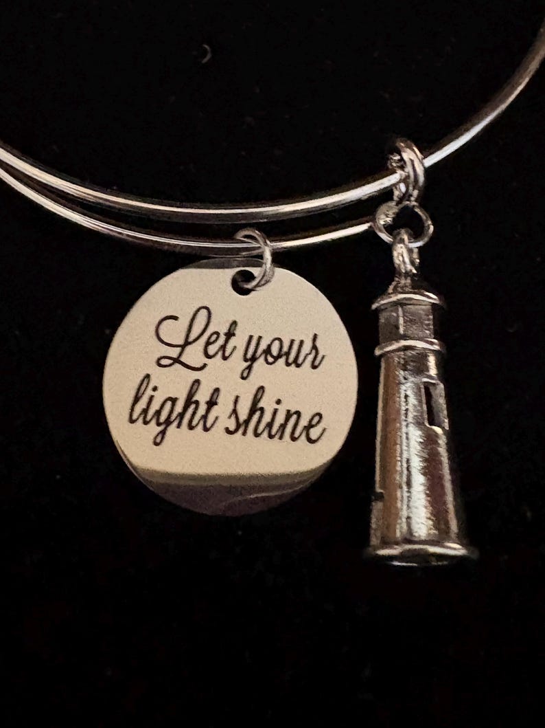 Puede incluir: Pulsera de brazalete plateada con un dije redondo y un dije de faro. El dije redondo tiene el texto "Let your light shine". El dije de faro tiene ventanas y un techo detallados.