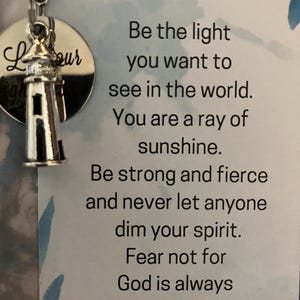 May include: A silver lighthouse charm with the word "Lour" on a round silver tag. The charm is attached to a white card with the text "Be the light you want to see in the world. You are a ray of sunshine. Be strong and fierce and never let anyone dim your spirit. Fear not for God is always within you."