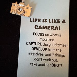 May include: A silver-colored camera charm hanging from a silver chain. The charm is next to a light blue card with the text "LIFE IS LIKE A CAMERA! FOCUS on what is important. CAPTURE the good times. DEVELOP from the negatives, and if things don't work out, take another SHOT!"