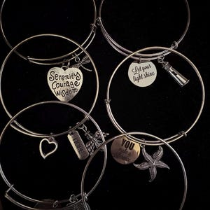 Puede incluir: Pulseras de brazalete plateadas con varios dijes. Los dijes incluyen un coraz&oacute;n con "Serenity Courage Wisdom", un faro, una estrella de mar y las palabras "Hope" y "You make a difference". Fondo negro.
