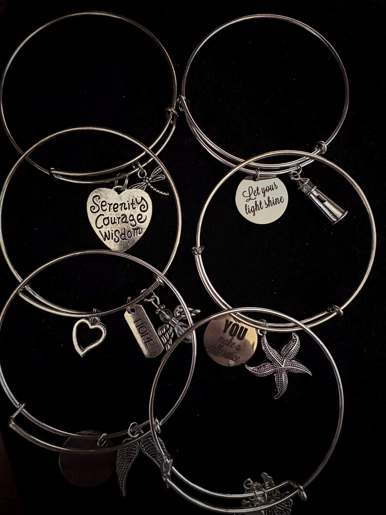 Puede incluir: Pulseras de brazalete plateadas con varios dijes. Los dijes incluyen un coraz&oacute;n con "Serenity Courage Wisdom", un faro, una estrella de mar y las palabras "Hope" y "You make a difference". Fondo negro.