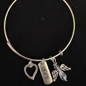 Puede incluir: Pulsera de brazalete plateada con tres colgantes. Los colgantes incluyen un coraz&oacute;n, una etiqueta rectangular con la palabra "HOPE" y un &aacute;ngel con detalles de cristal. La pulsera se muestra sobre un fondo negro.