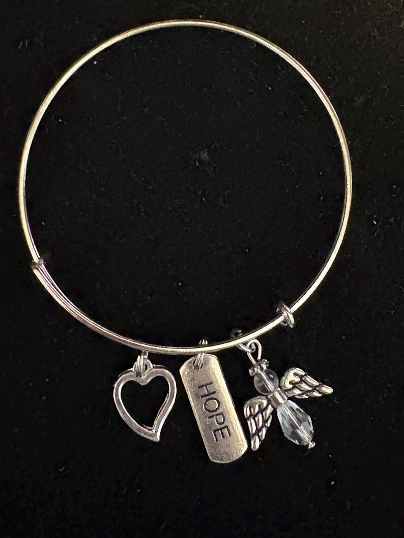 Puede incluir: Pulsera de brazalete plateada con tres colgantes. Los colgantes incluyen un coraz&oacute;n, una etiqueta rectangular con la palabra "HOPE" y un &aacute;ngel con detalles de cristal. La pulsera se muestra sobre un fondo negro.