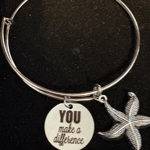 Puede incluir: Pulsera de brazalete plateada con un dije redondo y un colgante de estrella de mar. El dije est&aacute; grabado con las palabras "YOU make a difference". El colgante de estrella de mar tiene una textura detallada. La pulsera est&aacute; sobre un fondo oscuro.