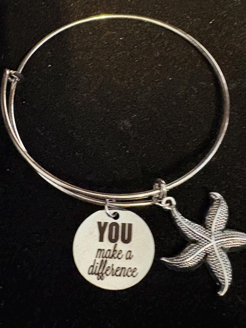 Puede incluir: Pulsera de brazalete plateada con un dije redondo y un colgante de estrella de mar. El dije est&aacute; grabado con las palabras "YOU make a difference". El colgante de estrella de mar tiene una textura detallada. La pulsera est&aacute; sobre un fondo oscuro.