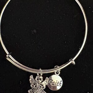 Puede incluir: Pulsera de brazalete plateada con dos colgantes. Uno es una figura de &aacute;ngel con alas detalladas, y el otro es un colgante redondo con la palabra "Faith". La pulsera est&aacute; sobre un fondo negro.
