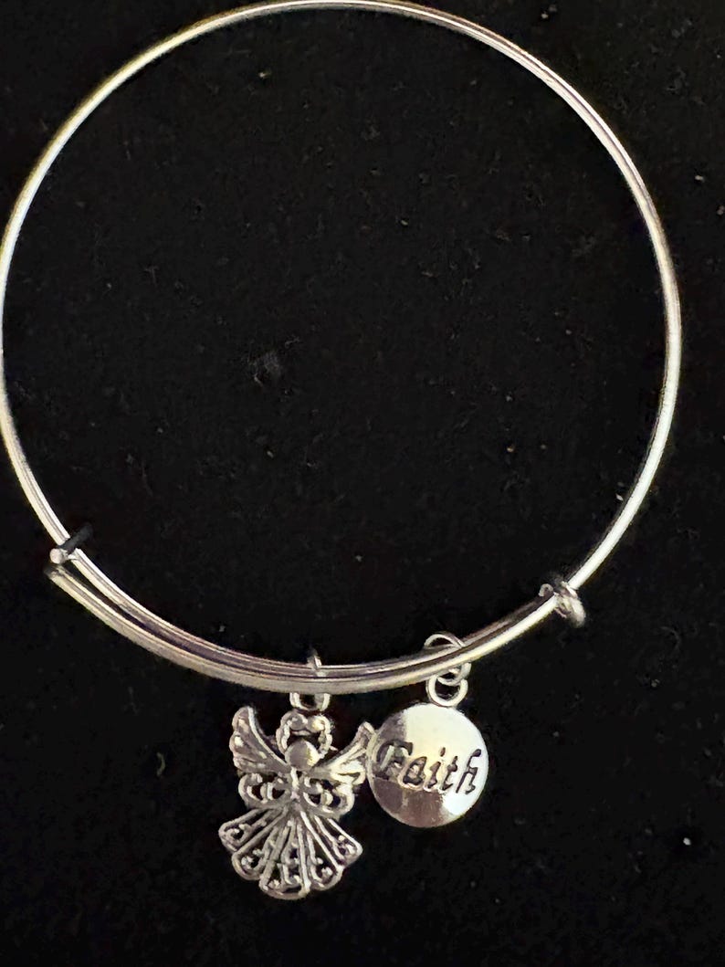 Puede incluir: Pulsera de brazalete plateada con dos colgantes. Uno es una figura de &aacute;ngel con alas detalladas, y el otro es un colgante redondo con la palabra "Faith". La pulsera est&aacute; sobre un fondo negro.