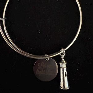 Puede incluir: Pulsera de brazalete plateada con un dije de faro y un dije redondo grabado con las palabras "Let your light shine". El dije de faro tiene ventanas y una luz. Sobre un fondo negro.