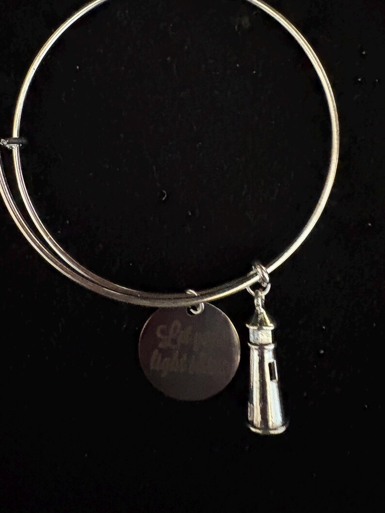 Puede incluir: Pulsera de brazalete plateada con un dije de faro y un dije redondo grabado con las palabras "Let your light shine". El dije de faro tiene ventanas y una luz. Sobre un fondo negro.