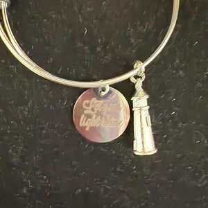 Puede incluir: Pulsera de brazalete plateada con dos colgantes. Uno es un disco redondo con las palabras "Let your light shine" grabadas. El otro es un faro en miniatura. La pulsera se muestra sobre un fondo oscuro.
