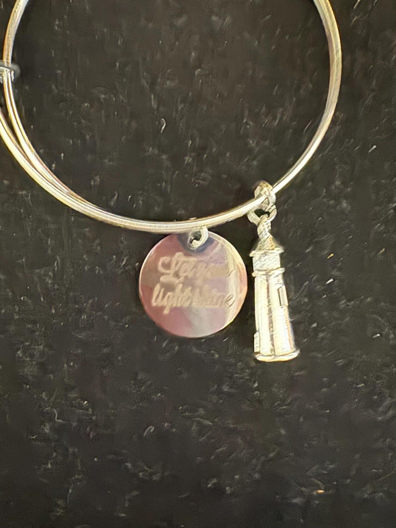 Puede incluir: Pulsera de brazalete plateada con dos colgantes. Uno es un disco redondo con las palabras "Let your light shine" grabadas. El otro es un faro en miniatura. La pulsera se muestra sobre un fondo oscuro.
