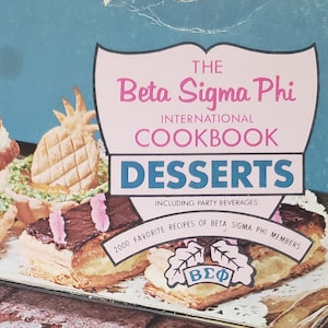 May include: A vintage cookbook titled "The Beta Sigma Phi International Cookbook: Desserts" with a blue cover and a photo of a layered cake with whipped cream and cherries on top.