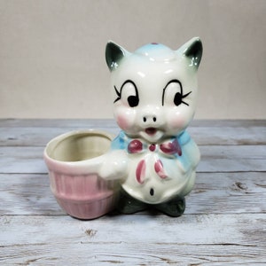 May include: A vintage ceramic pig-shaped planter. The pig is white with a light blue jacket and a pink bow tie. The planter is light pink and has a round shape. The planter is likely to be around 15 cm in height.