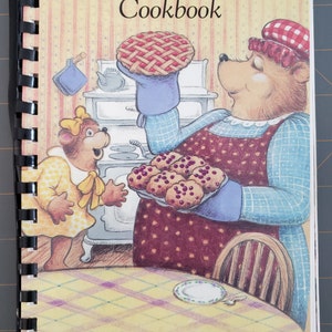 May include: A cookbook cover featuring two cartoon bears in a kitchen setting. The larger bear is holding a pie and a tray of muffins. The smaller bear is wearing a yellow dress and a bow. The text on the cover reads "Our 20th Anniversary Cookbook" and "Lambeth Co-operative Playschool Inc. Lambeth, Ontario."