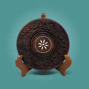 May include: A dark brown wooden coaster with intricate floral carvings and a white star-shaped inlay in the centre. The coaster is displayed on a wooden stand.