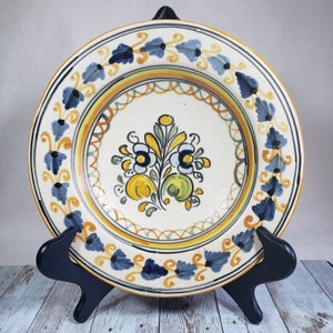 May include: A white ceramic plate with a blue and yellow floral design. The plate has a raised rim and a scalloped edge. It is decorated with a floral pattern in shades of blue, yellow, and green.