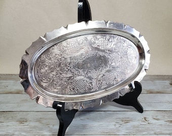 Puede incluir: Una bandeja de servir ovalada de plata con un diseño ornamentado y en relieve. La bandeja está sobre un soporte negro.