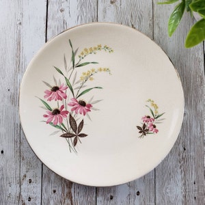 May include: A white ceramic plate with a gold rim features a floral design. Pink flowers with yellow centres are clustered on the left side of the plate. A smaller cluster of pink flowers is on the right side of the plate. The flowers are surrounded by green leaves and yellow flowers.