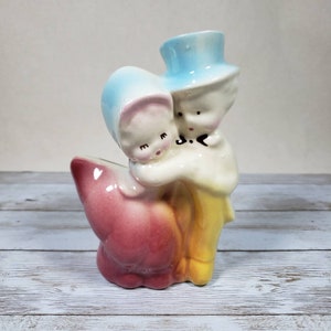 Peut inclure: Un pot de fleurs en céramique vintage en forme de couple enlacé. La femme porte un bonnet bleu et l'homme un chapeau haut de forme bleu. Le pot est peint dans des tons de rose, bleu, jaune et blanc.