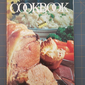 以下が含まれることがあります： 表紙にローストミート料理の写真が載っているビンテージの料理本。表紙のテキストは「it's mainly because of the meat」COOKBOOK。表紙に「2.59ドル」の値札が見える。