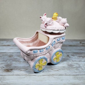 May include: A pink ceramic baby carriage with blue and yellow accents. The carriage has a removable top with three pig figurines and a yellow bird figurine. The carriage is a musical box.