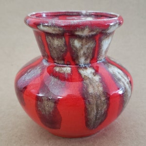 Op de afbeelding: Een kleine, rode keramische vaas met bruine en witte glazuur. De vaas heeft een rond lichaam en een uitlopende opening.