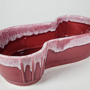 Peut inclure: Un pot en céramique rouge avec un glaçage blanc, en forme de long ovale avec un bord incurvé. Le pot a un effet de glaçage en goutte.