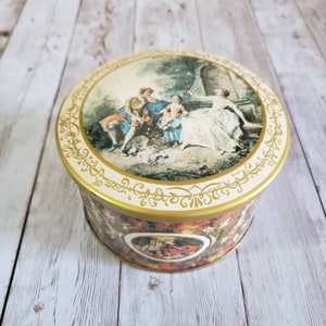 Op de afbeelding: Een ronde vintage blikken doos met een gouden rand en een kleurrijk beeld van een stel in een tuin. Het deksel heeft een gouden bloemmotief.