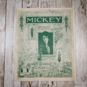 May include: A vintage sheet music cover for the song "Mickey" with a green background and a photo of Mabel Normand. The text on the cover reads "Adapted from Mack Sennett's 500,000 Photoplay "Mickey" and respectfully dedicated to Miss Mabel Normand."