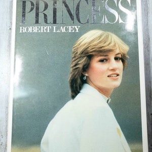 Puede incluir: Portada de libro con el título "PRINCESS" en letras plateadas y el nombre del autor, Robert Lacey. La portada muestra un retrato de una mujer con cabello rubio y una chaqueta blanca sobre un fondo gris.
