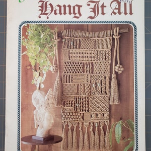 May include: A book titled "Juliano's Hang It All" featuring a brown macrame wall hanging with tassels and beads. The book cover has a wood-toned background and a rope border.
