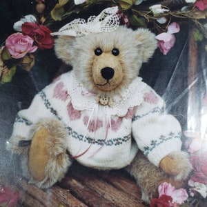May include: A plush teddy bear wearing a white sweater with pink hearts and a white lace collar. The bear is sitting on a wooden surface with pink flowers in the background.