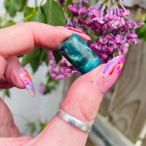 Könnte beinhalten: Ein rechteckiger Kristall in Grün und Blau mit Farbwirbeln. Der Kristall wird von einer Hand mit buntem Nagellack gehalten.
