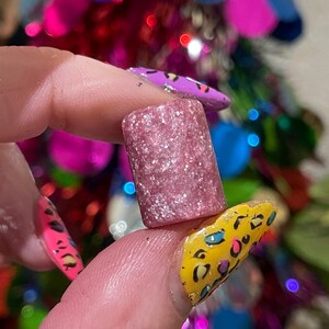 Könnte beinhalten: Ein rosa Glitzerzylinder mit weißem Schimmer. Der Zylinder wird zwischen zwei Fingern gehalten.