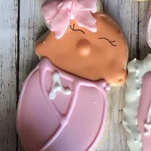 Puede incluir: Una galleta de azúcar rosa y naranja decorada con una niña bebé en un mono rosa y un lazo rosa. La galleta tiene una letra 'A' blanca en el mono.