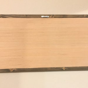 May include: Back of a rectangular wooden sign with a light brown wood panel and a dark brown wooden frame. A metal hanger is attached to the top center of the frame. The sign is mounted on a white wall.