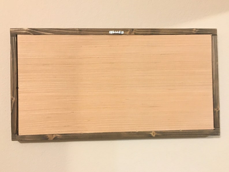 May include: Back of a rectangular wooden sign with a light brown wood panel and a dark brown wooden frame. A metal hanger is attached to the top center of the frame. The sign is mounted on a white wall.