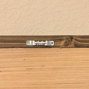 May include: Close-up of the back of a wooden framed artwork. The frame is made of dark brown stained wood. A metal sawtooth hanger is attached to the top of the frame for hanging on a wall. The artwork is made of light brown wood.