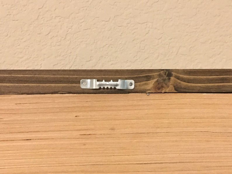 May include: Close-up of the back of a wooden framed artwork. The frame is made of dark brown stained wood. A metal sawtooth hanger is attached to the top of the frame for hanging on a wall. The artwork is made of light brown wood.