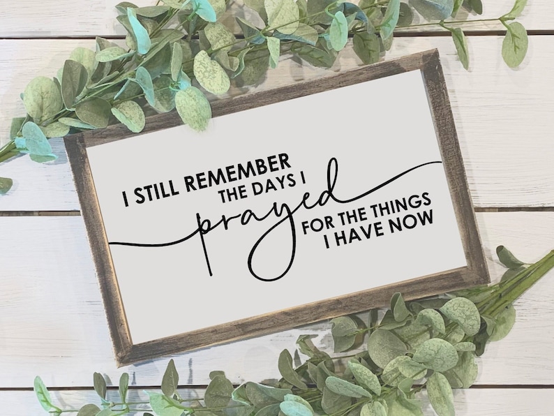May include: A framed sign with the text "I still remember the days I prayed for the things I have now" in black script. The sign has a white background and a rustic, brown wooden frame. Greenery surrounds the sign.