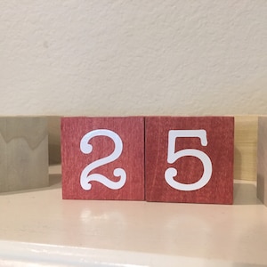 May include: Wooden blocks displaying the number 25 in white against a red backdrop. The blocks are positioned on a white surface, with additional wooden blocks visible. These blocks are likely part of a decorative calendar.