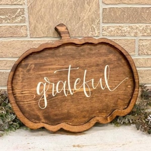 Puede incluir: Un letrero de madera con forma de calabaza con la palabra "agradecido" escrita en pintura blanca.
