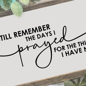 May include: A rectangular wooden sign with the quote, "I still remember the days I prayed for the things I have now," in black script. The sign has a light background and a dark brown frame, with greenery in the corners.