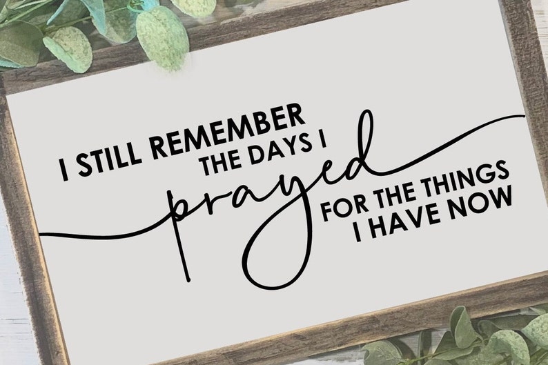 May include: A rectangular wooden sign with the quote, "I still remember the days I prayed for the things I have now," in black script. The sign has a light background and a dark brown frame, with greenery in the corners.