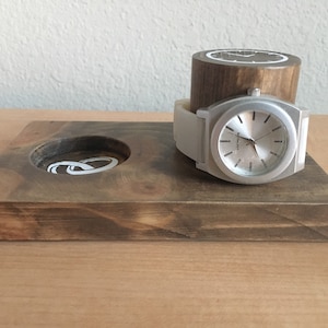 May include: A wooden watch stand with a watch and a ring holder. The stand is made of dark brown wood and has a natural finish. The watch is silver with a white face and a white band. The ring holder is a small circle in the center of the stand.